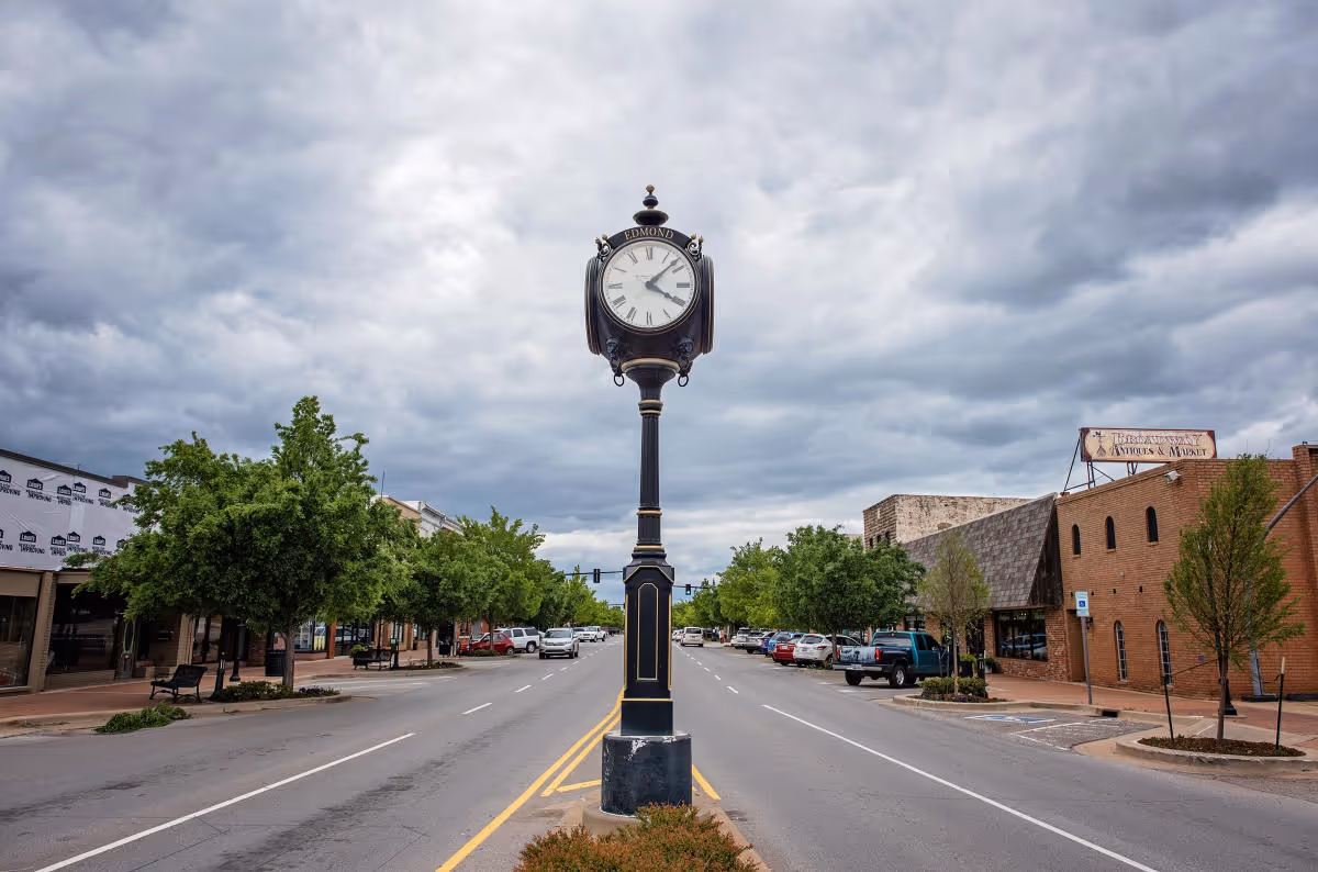 Black street clock labeled Edmond standing on a divider in the middle of a two-way street lined with trees and parked cars under a cloudy sky.