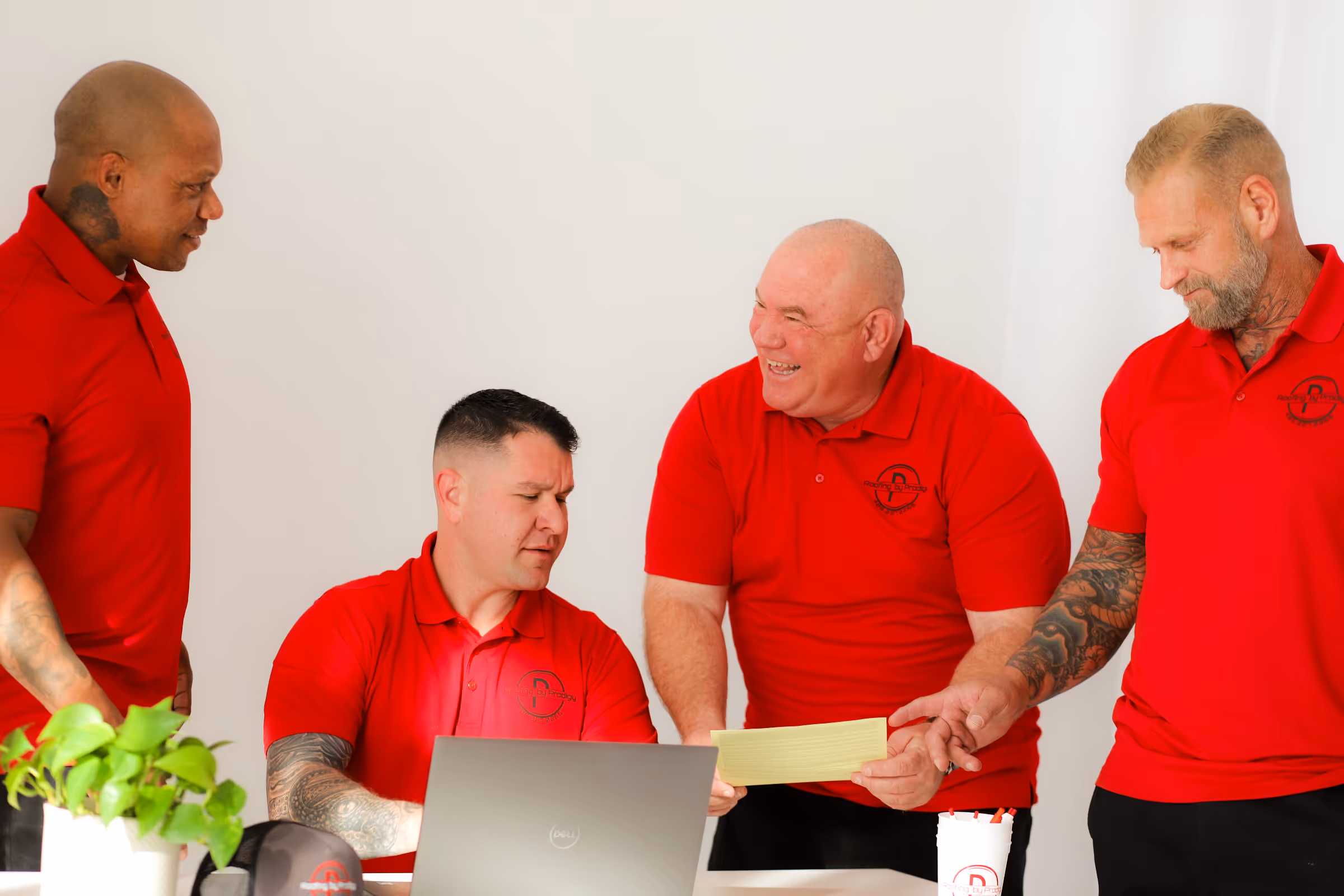 Roofing by Prodigy - Four men in red polo shirts collaborating around a laptop and a yellow notepad in a bright room.
