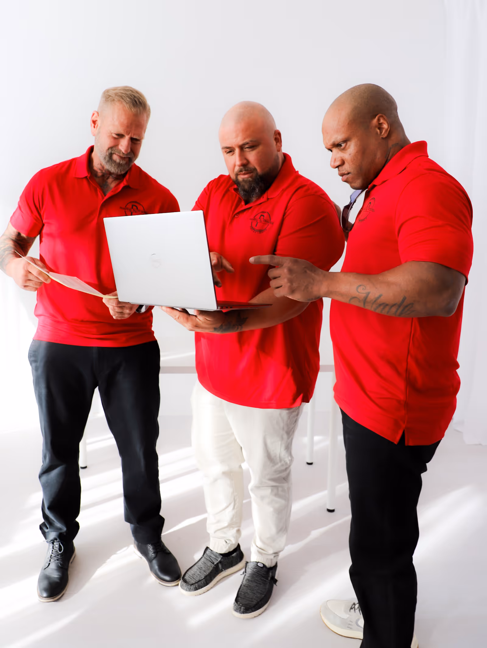 Three men in red polo shirts looking at a laptop and a piece of paper in a bright, white room.