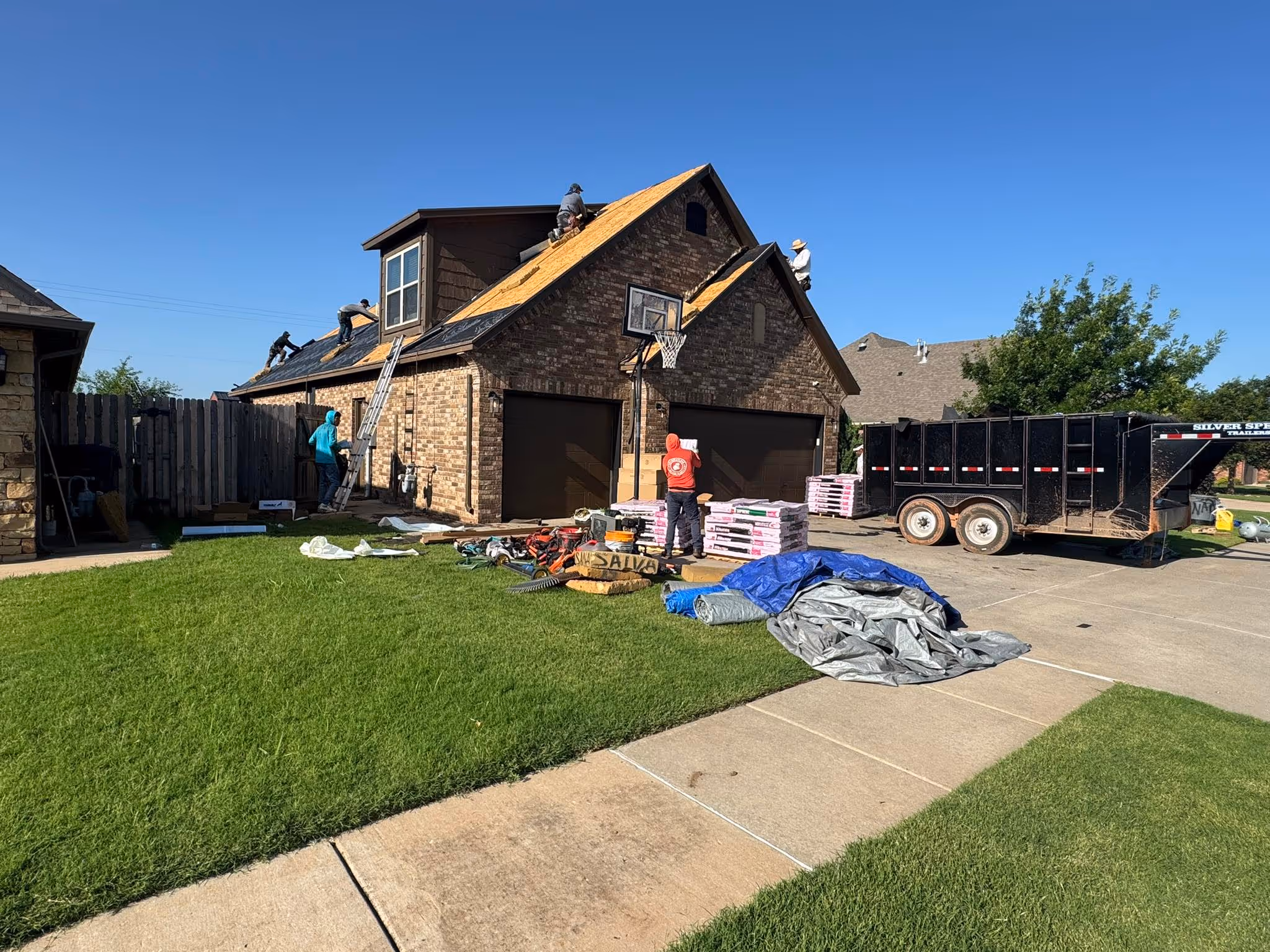 Crew replacing the roof on a two-story stone home, with materials and a dump trailer in the driveway.