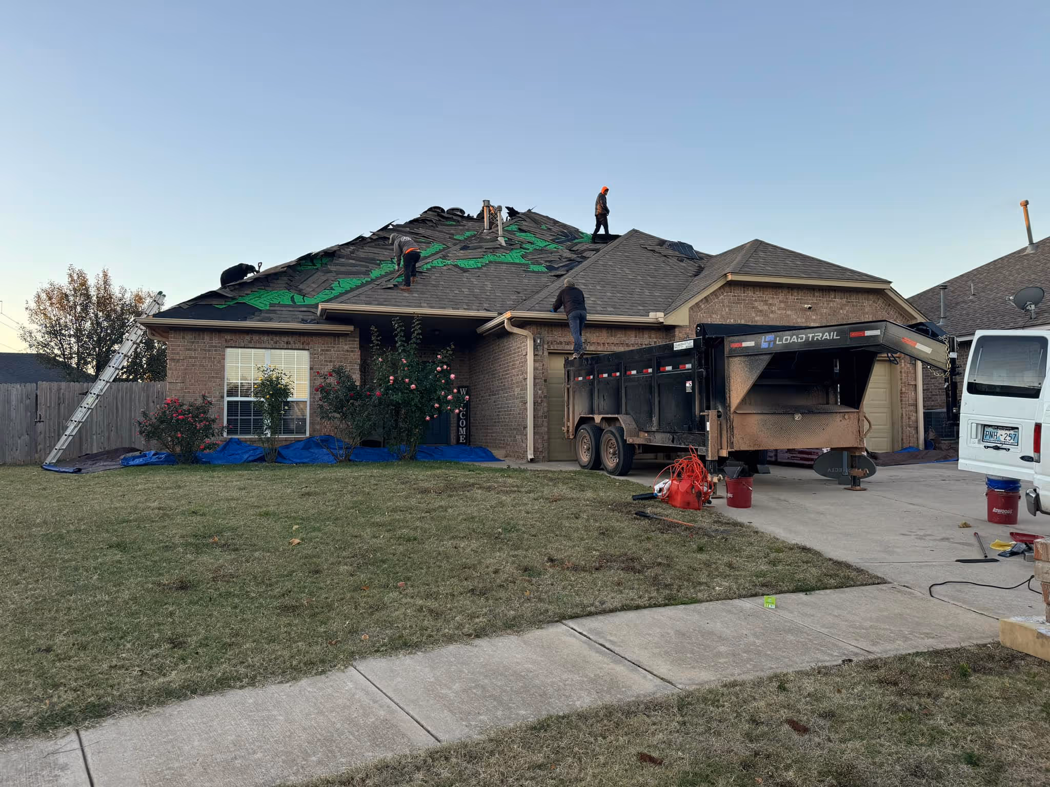 Roof replacement underway on a brick home, with new underlayment going down and a crew trailer in the driveway.