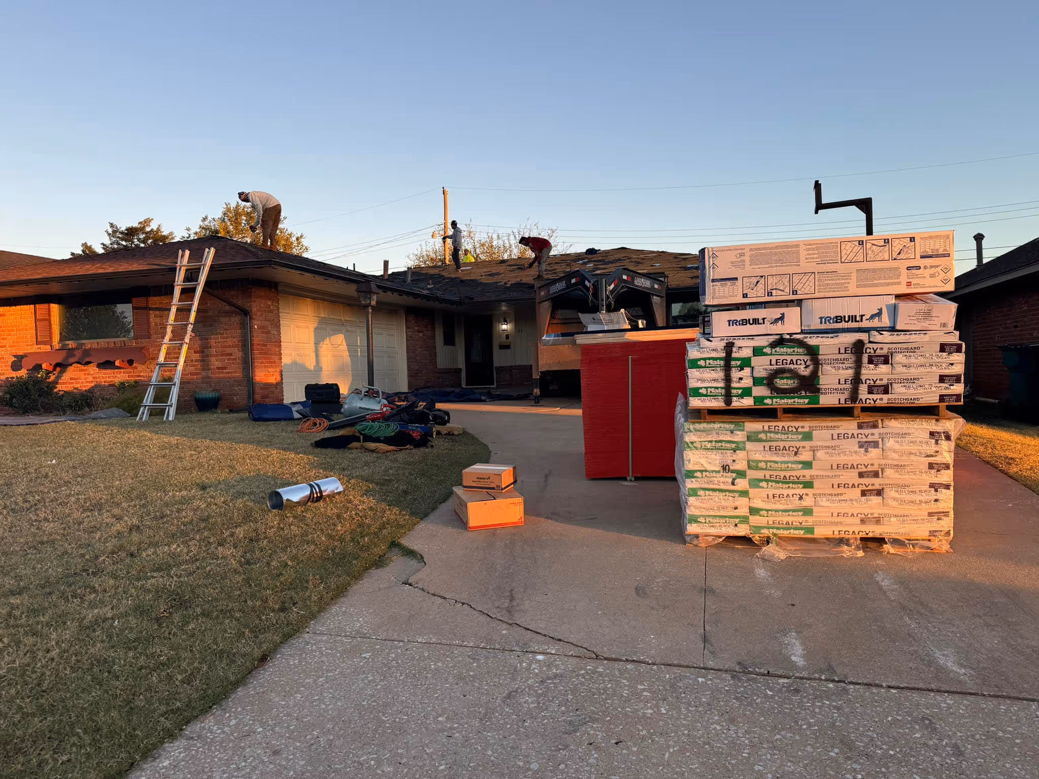 Three workers on a roof doing repairs with roofing materials stacked on the driveway in front of a brick house during a beautiful Oklahoma sunset.