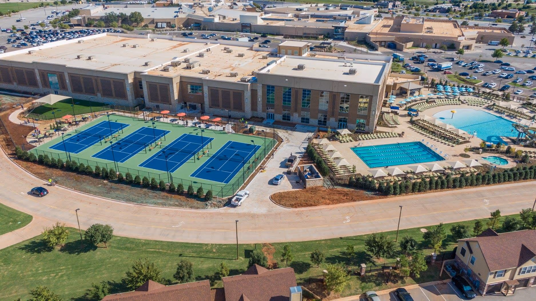 Aerial view of a large recreational complex featuring multiple tennis courts, a swimming pool, a children's splash pool, lounge chairs with umbrellas, a hot tub, surrounding greenery, and adjacent parking lots.