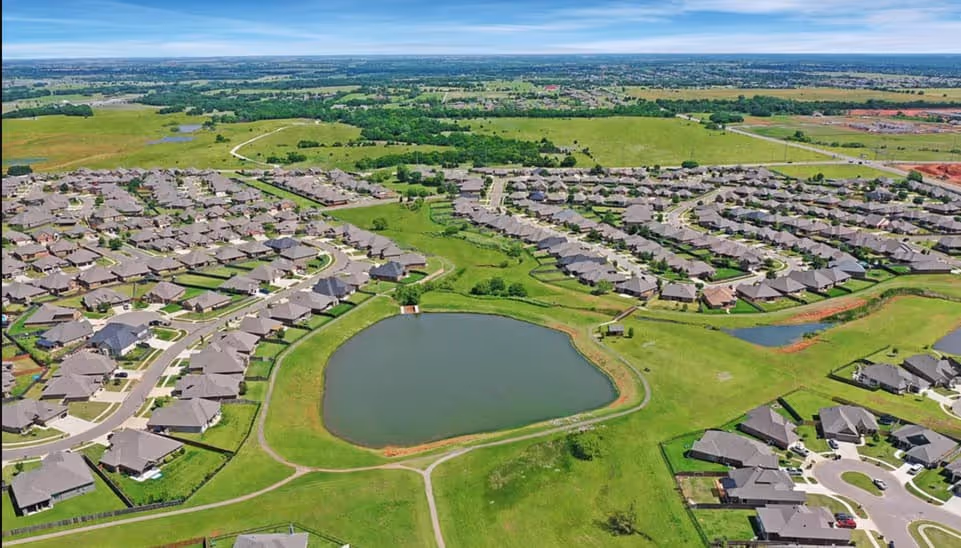 Aerial view of a suburban neighborhood surrounding a central pond with green lawns and winding streets.