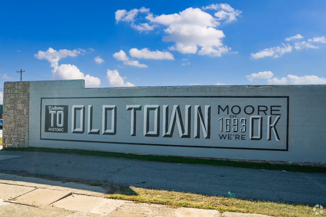 Mural on a wall reading 'Welcome to Historic Old Town Moore EST. 1893 We're OK' under a blue sky with clouds.